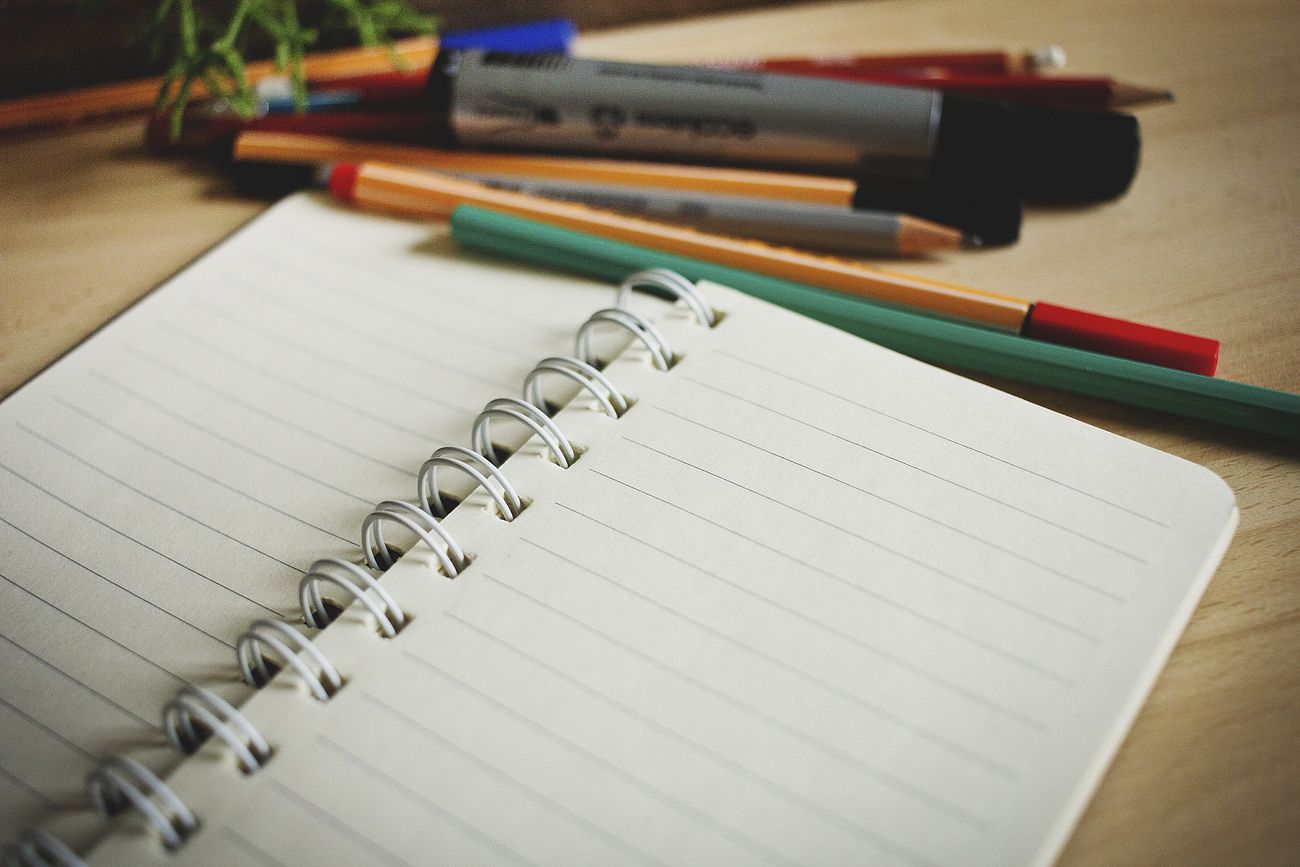 Image of an open spiral bound notebook with empty pages, surrounded by different pens, pencils, and a marker.
